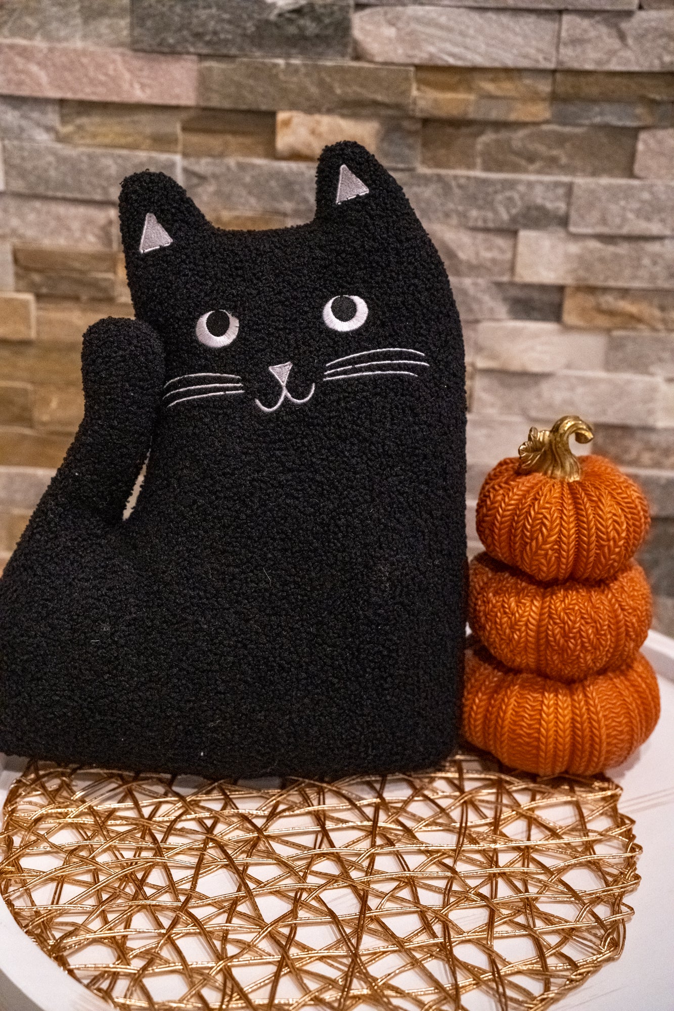 Knitted black cat with a small stack of orange pumpkins against a stone wall background