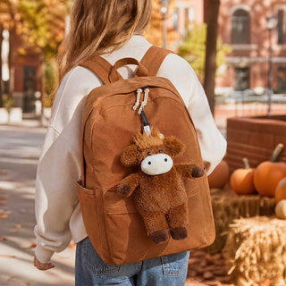 Warmies Highland Cow plush bag charm clipped to a brown canvas backpack outdoors