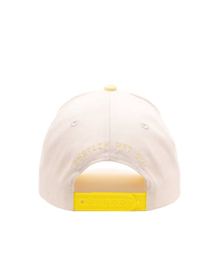 The back of a light cream baseball cap with 'RUSTLED HAT CO' embroidered in cream on the back panel and a bright yellow plastic snapback closure.