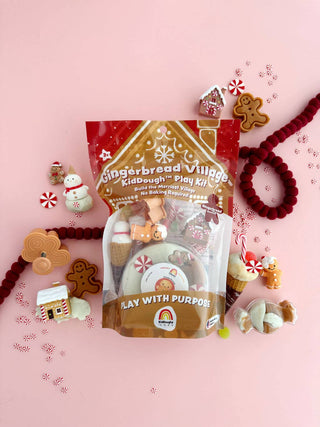 Gingerbread Village KidDough Play Kit bag surrounded by holiday figures and peppermint candies on pink
