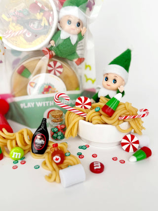 Elf dolls playing in a bowl of dough noodles with M&Ms, candy canes, and Hershey's syrup