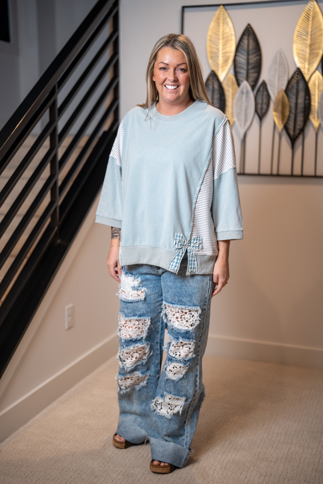 Woman wearing a light blue easel top and distressed jeans by oli & hali standing in a room with decorative wall art.