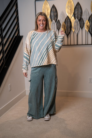 Woman wearing a patterned sweater by oli & hali and teal easel pants standing in a room with decorative wall art.