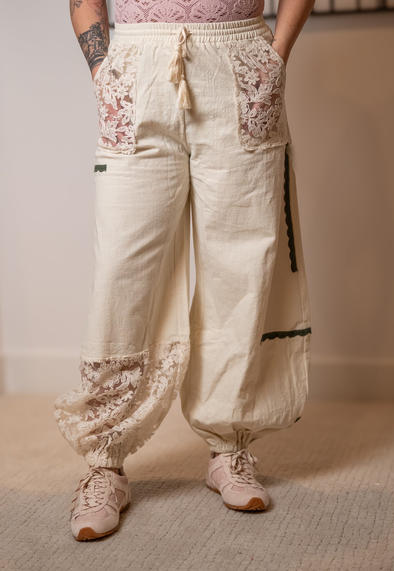 Person wearing cream-colored pants with lace detailing and sneakers on a neutral background