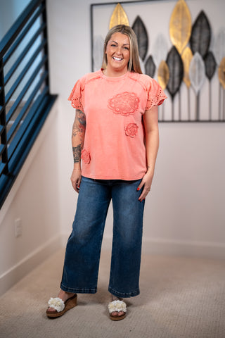 Woman wearing a pink t-shirt and blue jeans standing indoors with decorative wall art in the background.