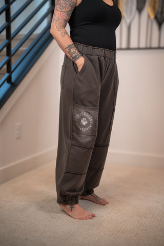 Person wearing brown cargo pants with a logo, standing in a room with a white wall and blue railing.