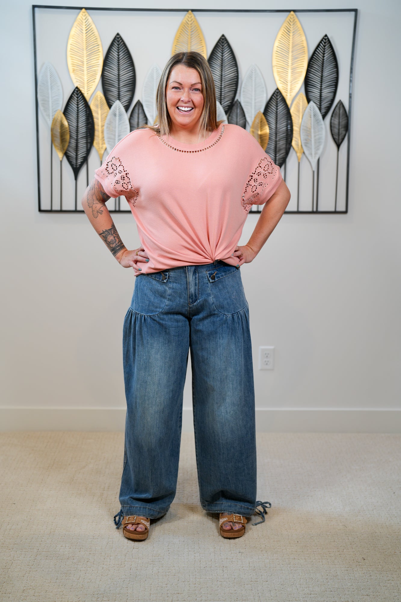 Woman wearing a pink top and blue jeans standing in a room with decorative wall art.