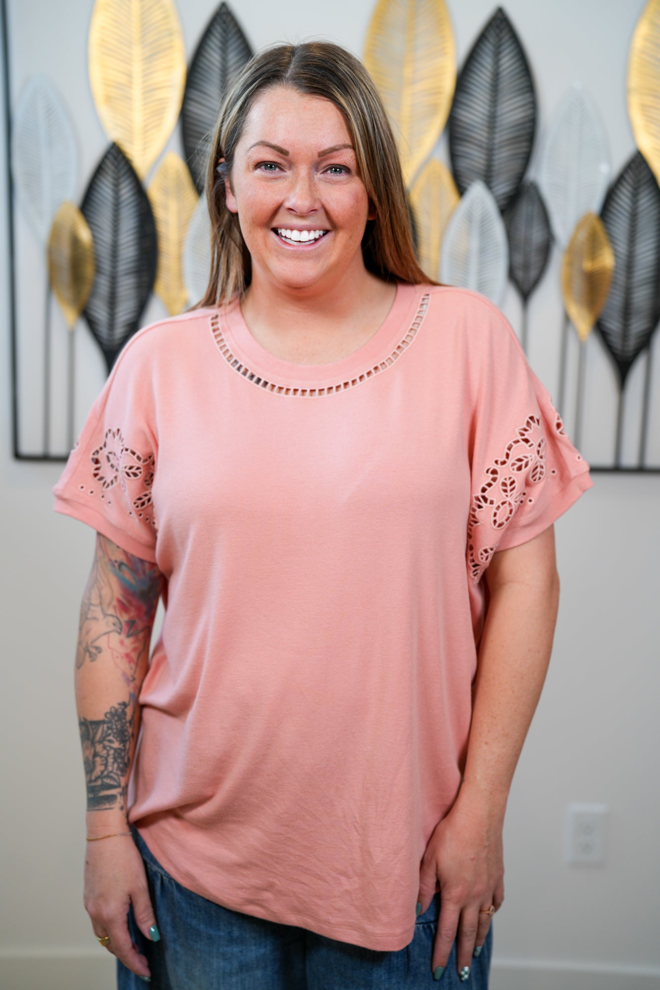 Woman wearing a pink blouse with lace sleeves in front of decorative wall art.