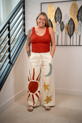 Woman wearing a red top and white pants with sun and moon designs, standing in a home setting.