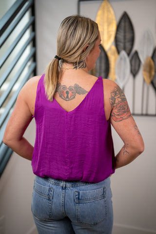 Person wearing a purple tank top and blue jeans with tattoos visible, standing in front of a decorative wall.