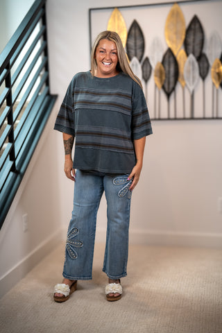 A woman stands indoors modeling a loose short-sleeve top with horizontal dark gray and black stripes, paired with light blue jeans featuring decorative embroidered details near the lower legs, and white fuzzy slide sandals.