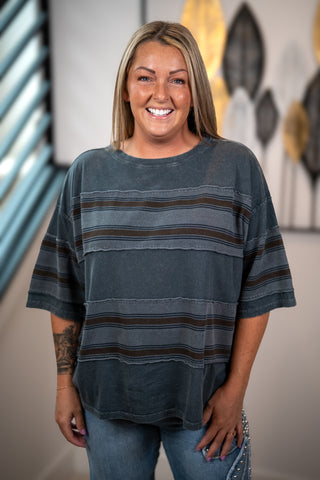 A smiling woman with blonde hair wearing a loose-fitting, charcoal gray short-sleeve t-shirt with horizontal brown and gray stripes, paired with blue jeans.