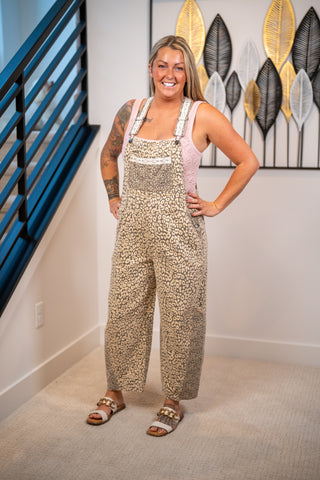 A woman modeling a beige and brown leopard print overall jumpsuit with a light pink underlayer and white straps, standing in a home setting with a staircase and wall art in the background.