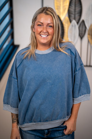 A smiling blonde woman wearing an oversized washed blue sweatshirt with gray ribbed trim, paired with blue jeans, standing indoors against a wall with decorative wall art.