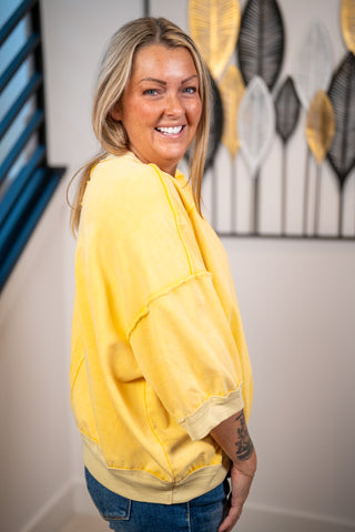 A smiling blonde woman wearing a bright yellow oversized sweatshirt and blue jeans, standing indoors against a wall with decorative wall art.