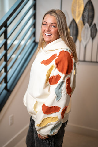 Cream hoodie with colorful abstract frayed fabric patches in rust, blue, and yellow
