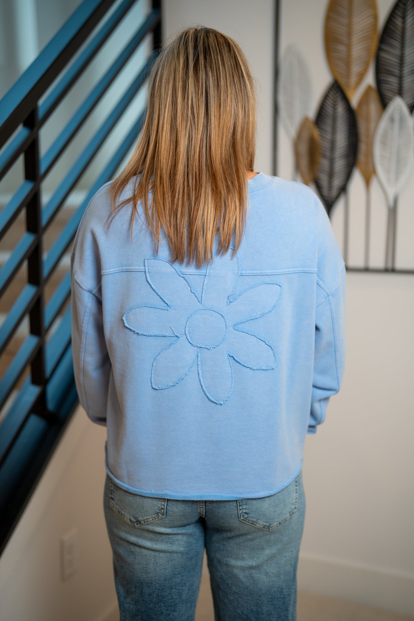 Person wearing a light blue sweatshirt with a floral design, standing in a room with a decorative wall art piece.