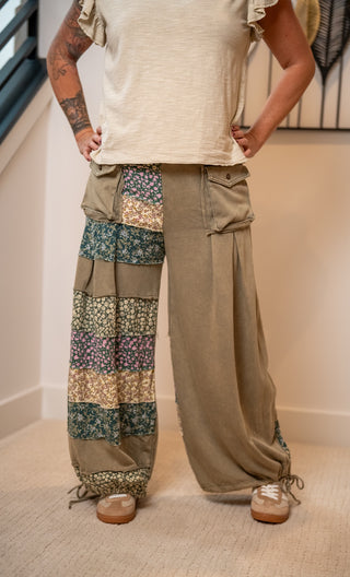 A person wearing wide-leg patchwork cargo pants in a beige and multicolor floral print, paired with a cream t-shirt and brown shoes, standing indoors.