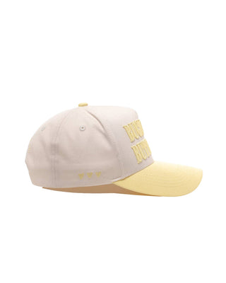 A baseball cap in off-white with a pale yellow brim and matching pale yellow embroidered lettering on the side, shown against a plain white background.