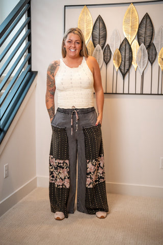 A woman stands indoors modeling a sleeveless white knit top and wide-leg patchwork pants with floral and dotted patterns. The setting includes a staircase railing on the left and a decorative wall art piece behind her.